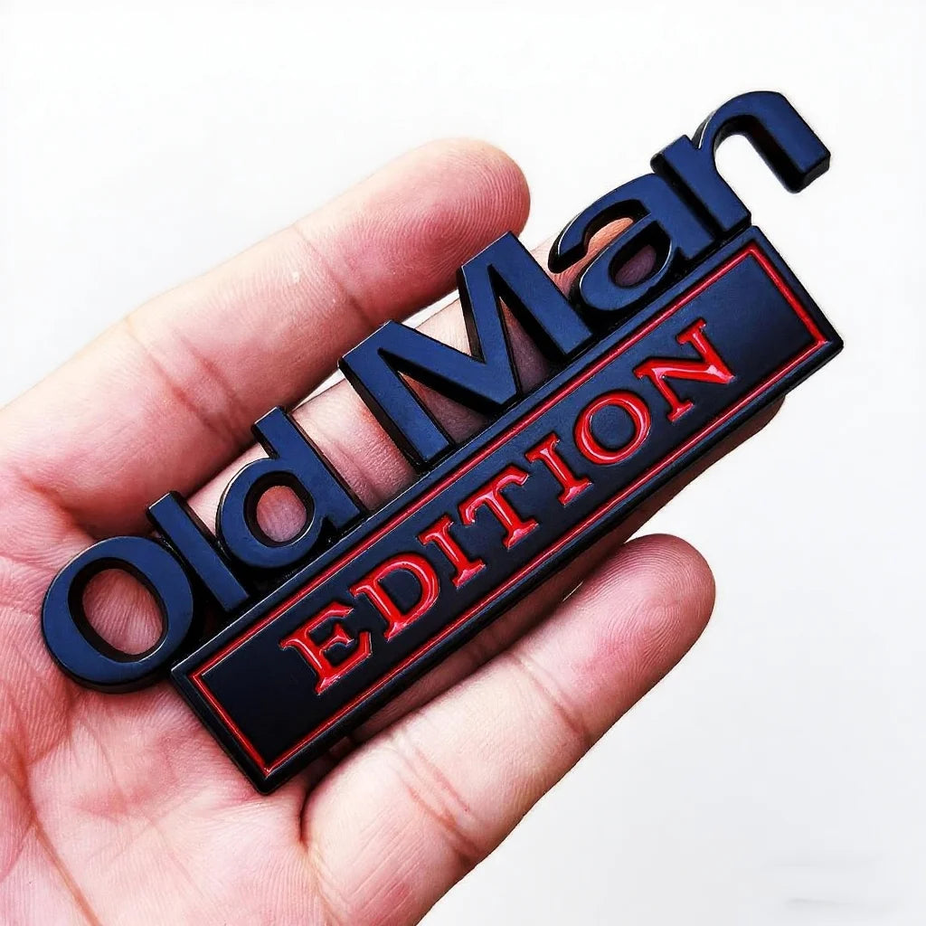 A hand holds a black and red Old Man Edition emblem badge set for a car fender.