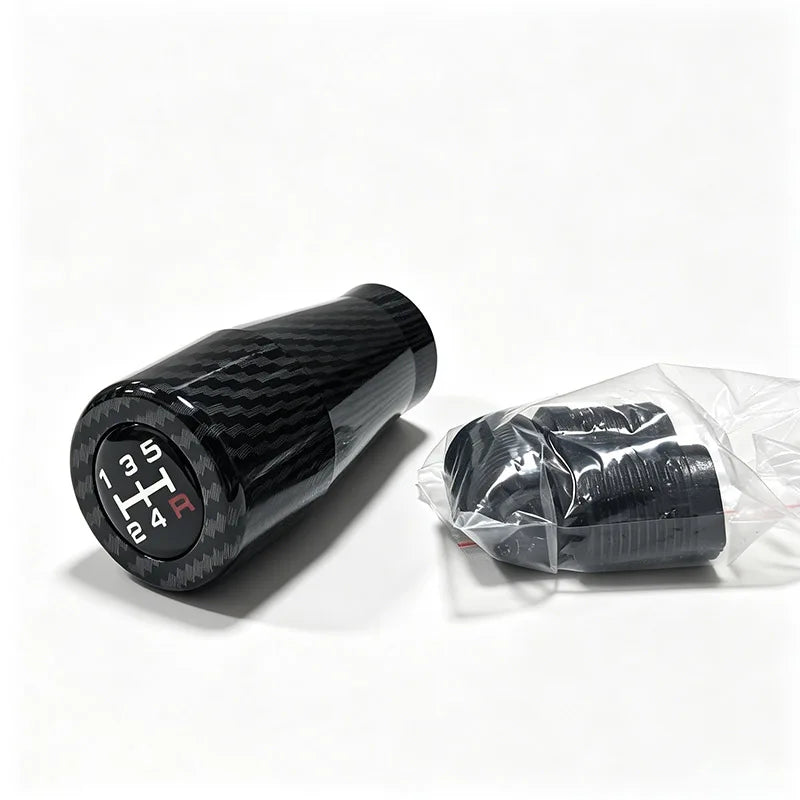 A black gear shift knob with a carbon fiber pattern is shown next to a clear plastic bag containing several knobs.