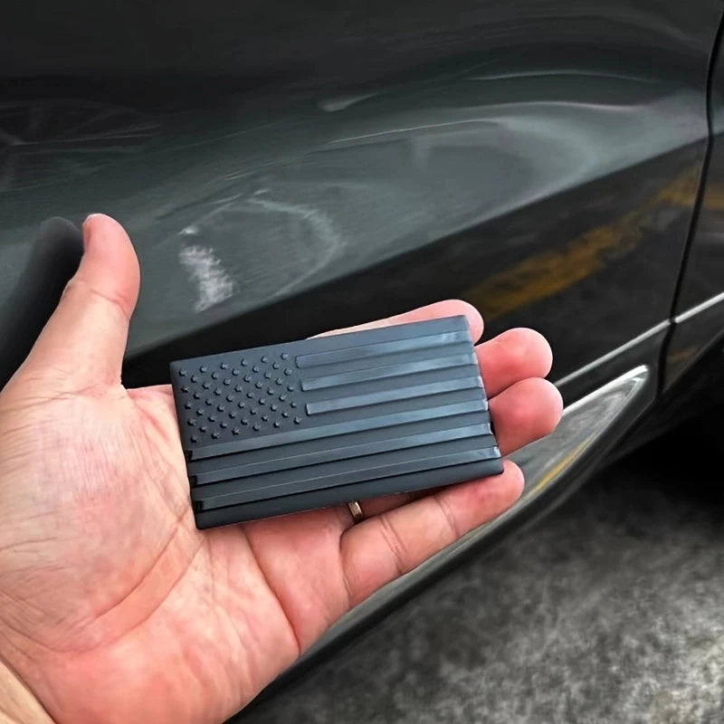 A hand holds a matte black 3D American flag decal for vehicles, featuring stars and stripes.