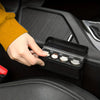 A hand is opening a black coin storage box with several coins inside, resting on a car interior surface.