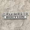 A silver metal emblem with the text SEMPER FI EDITION is displayed on a textured gray surface.