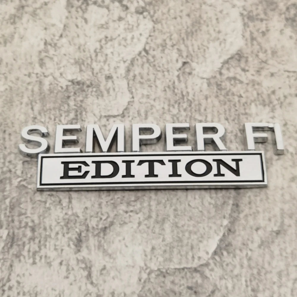 A silver metal emblem with the text SEMPER FI EDITION is displayed on a textured gray surface.