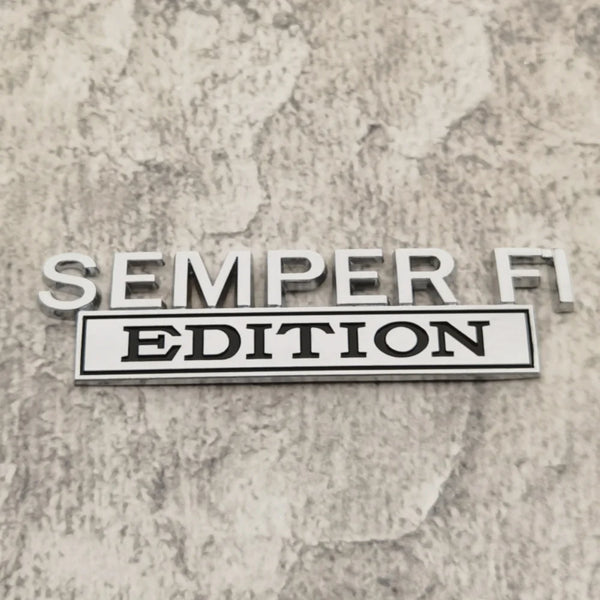 A silver metal emblem with the text SEMPER FI EDITION is displayed on a textured gray surface.