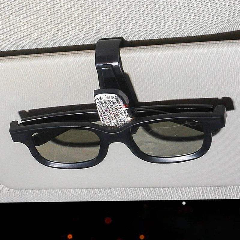 Eine schwarze Sonnenbrille liegt in einem Halter mit Kristallakzent auf einer hellen Oberfläche.