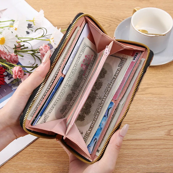 A hand holds an open pink long wallet with a gold zipper, displaying various banknotes and cards on a wooden surface.