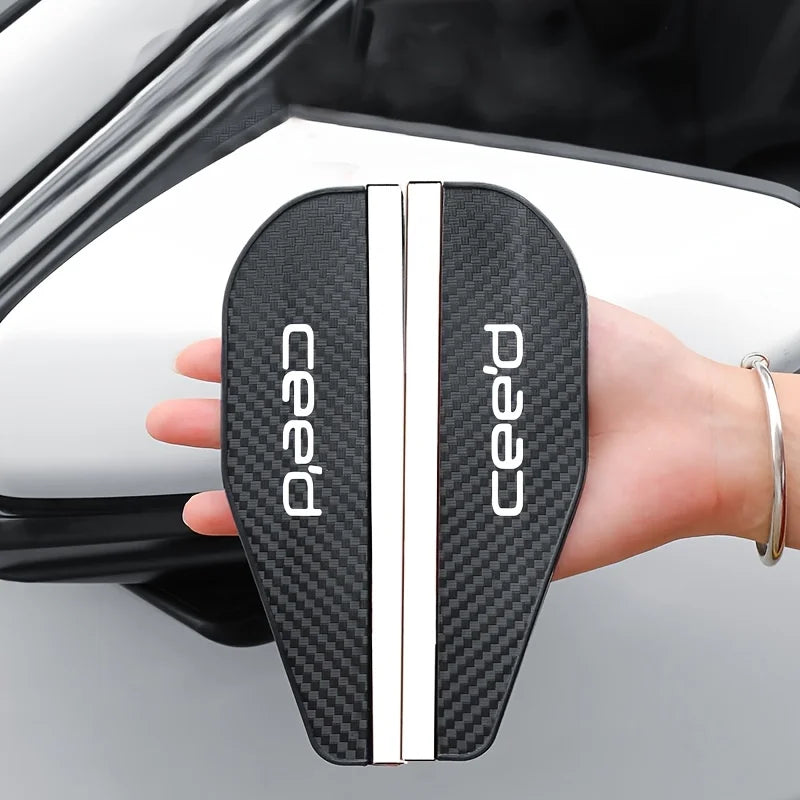 A hand holds a black carbon fiber rain shield designed for a car's rearview mirror.