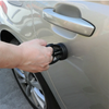 A hand is using a suction cup dent puller tool on the door handle of a light-colored car.