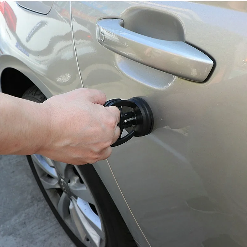 A hand is using a suction cup dent puller tool on the door handle of a light-colored car.