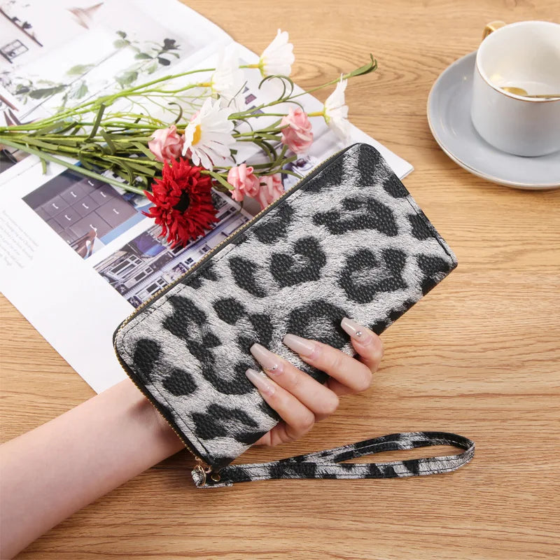 A hand holds a black and white leopard print long wallet on a wooden table next to some flowers and papers.