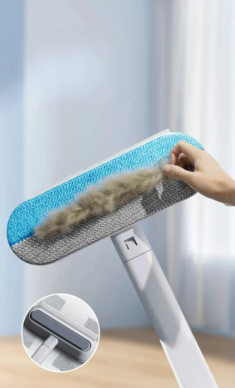 A hand is using a long-handled static pet hair remover brush with blue and gray bristles to clean a surface.