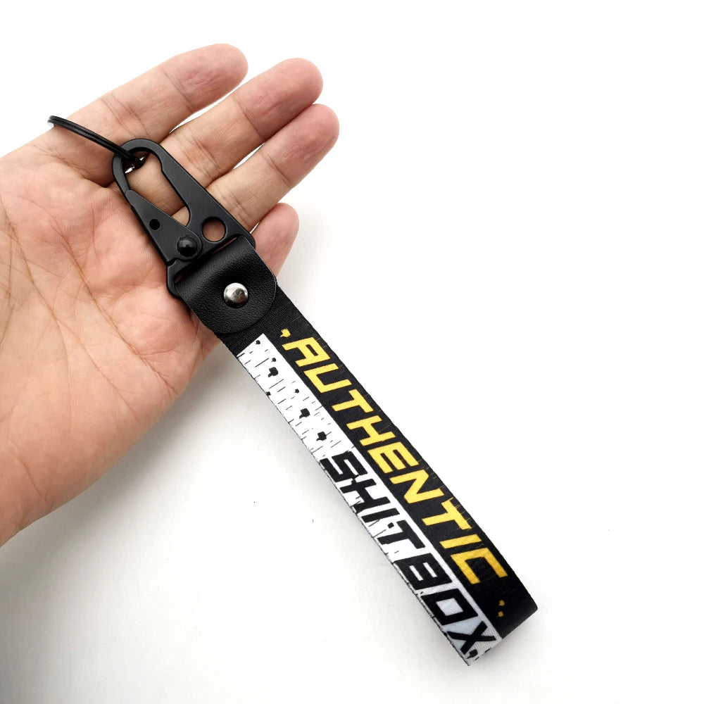 Ein schwarzer Lanyard mit weißer Schrift, die AUTHENTIC SHITBOX buchstabiert, wird in einer Hand gehalten.