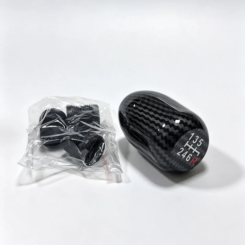 A black gear shift knob is shown next to a clear plastic bag containing a black manual gear shift knob.