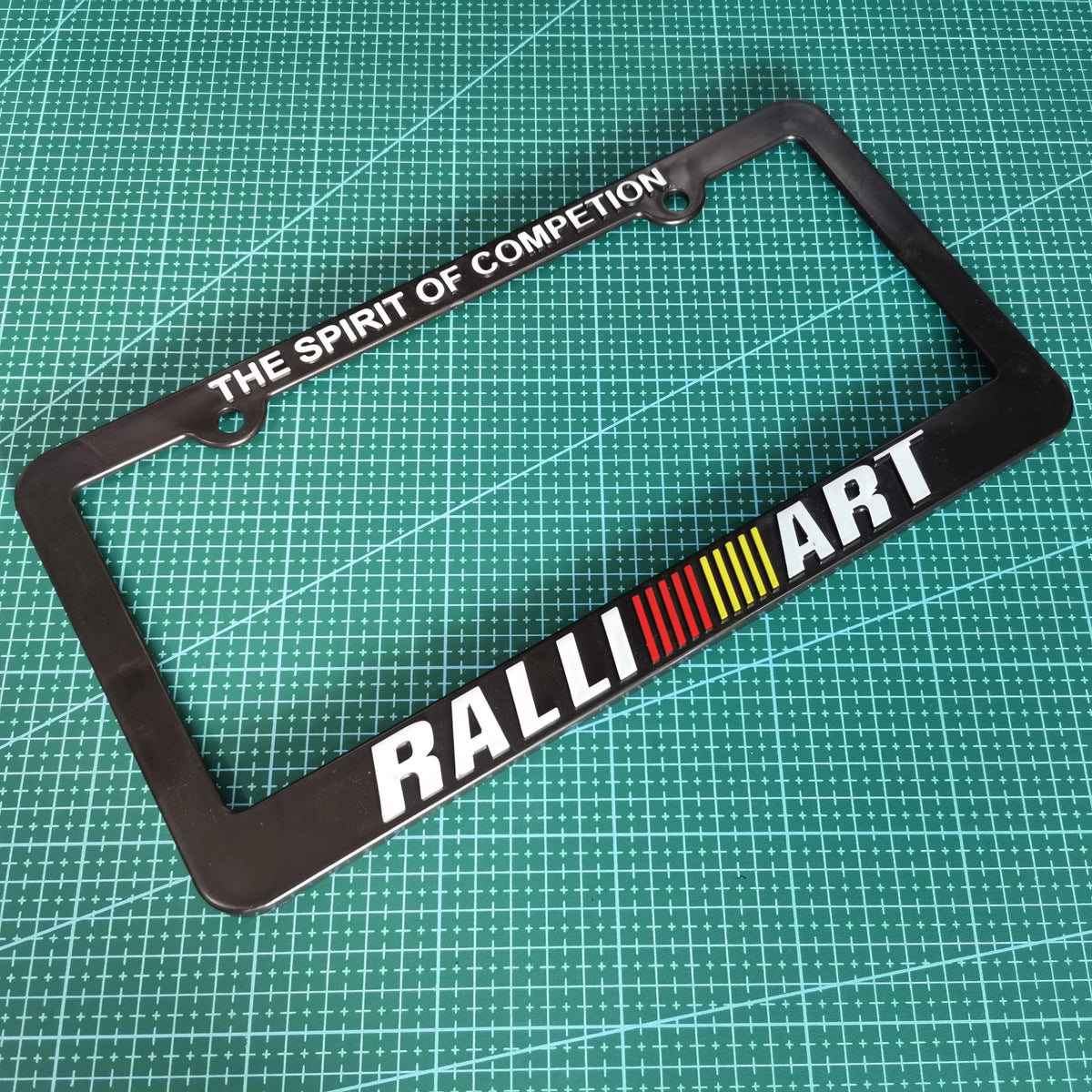 Black license plate frame decoration with the text THE SPIRIT OF COMPETITION and RALLY ART on it.