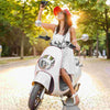 A woman wearing sunglasses and an orange hat is riding a white scooter with decorative trimming tape.