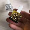 A hand holds a gold-colored car air freshener bottle with a white cap, next to a silver version.