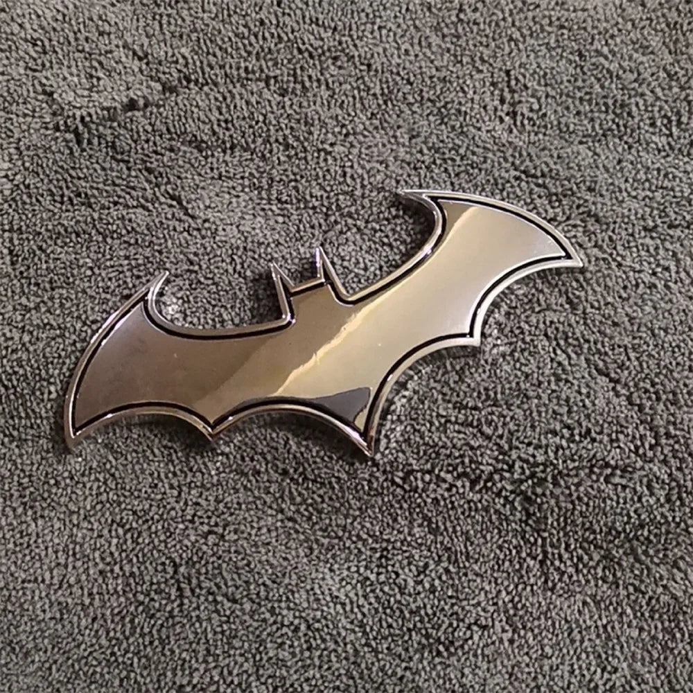 Silberfarbiger 3D-Metall-Bat-Aufkleber-Abzeichen auf einer dunkelgrauen, strukturierten Oberfläche.