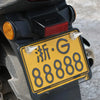 A close-up of a yellow license plate with black characters and silver screws mounted on a dark vehicle body.