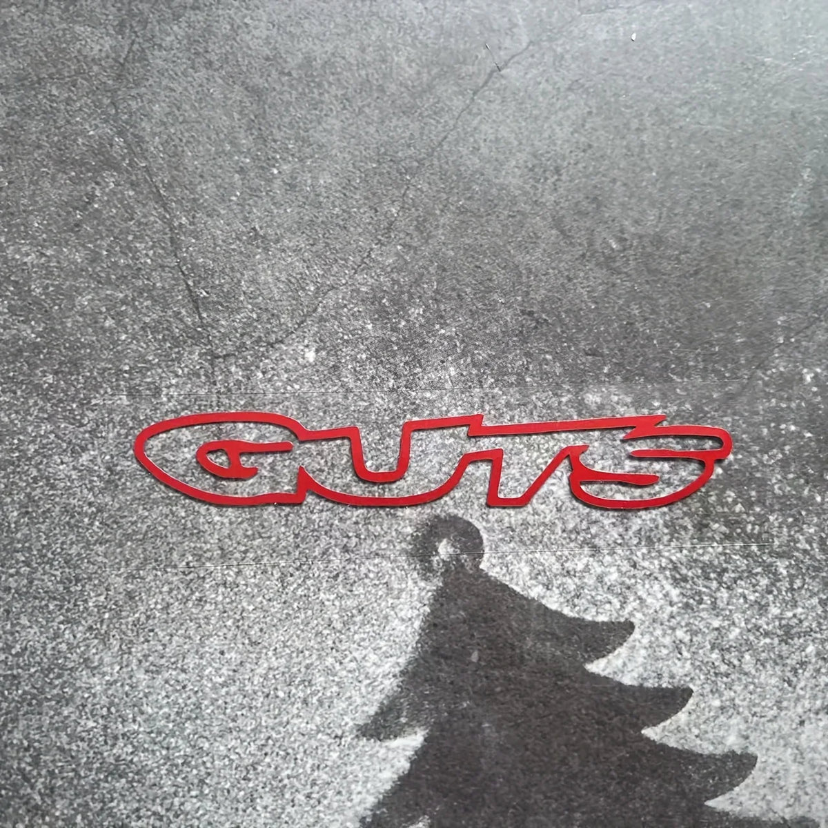 A red silhouette of the word GUTS is reflected on a wet, dark surface.