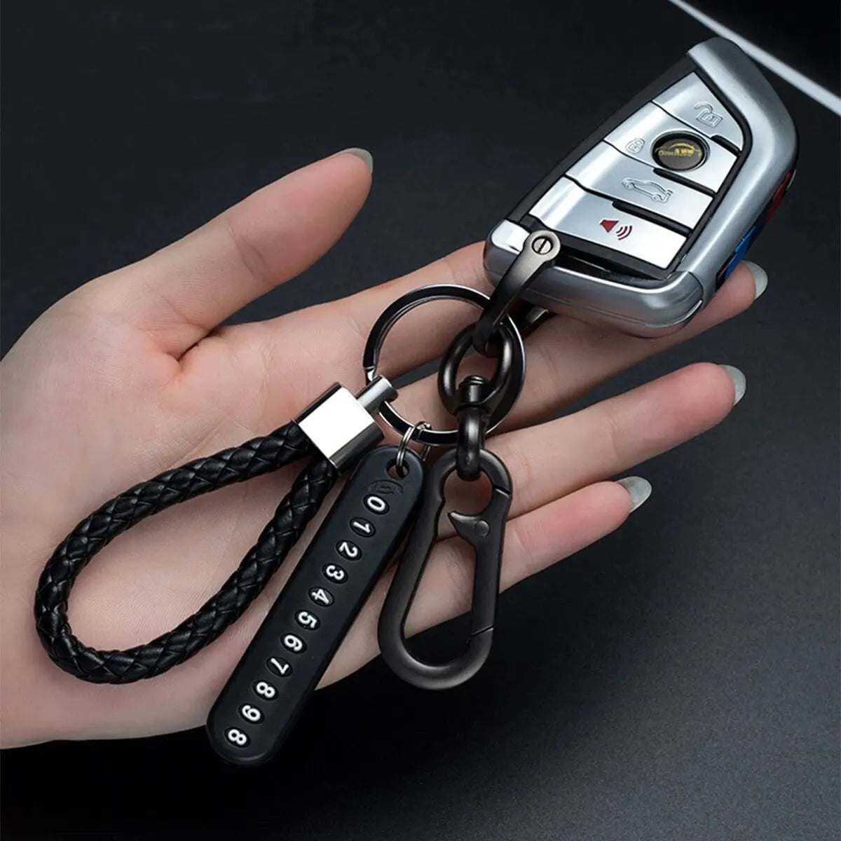 Eine Hand hält eine silberne Telefonnummer-Schlüsselanhänger mit schwarzem Band und einer Metallverschlusspfanne.