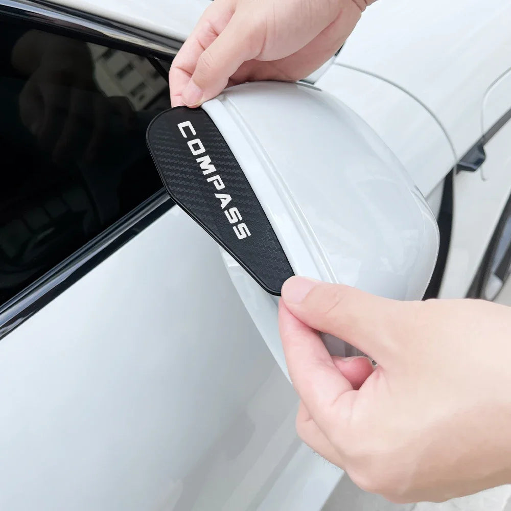 A hand is installing a black carbon fiber texture rain visor onto a white car's rearview mirror.