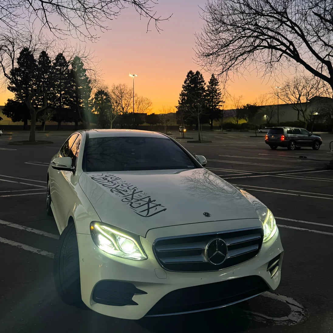 Une berline Mercedes-Benz blanche garée dans un parking au coucher du soleil avec des autocollants en écriture arabe sur le c