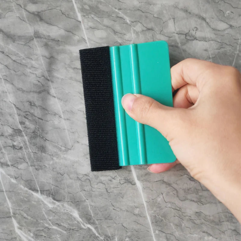 A hand holds a black felt squeegee tool with a teal handle against a gray marble background.