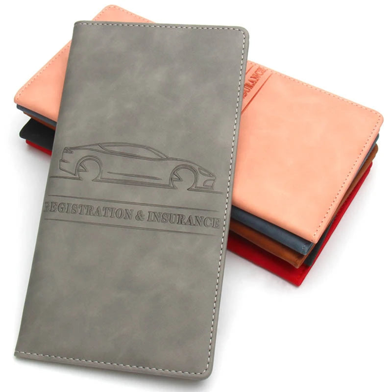 A grey leather document holder with a silhouette of a car embossed on the cover, shown next to other colored leather folders.