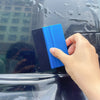 A hand is using a blue and black automotive film installation tool on a dark car window.