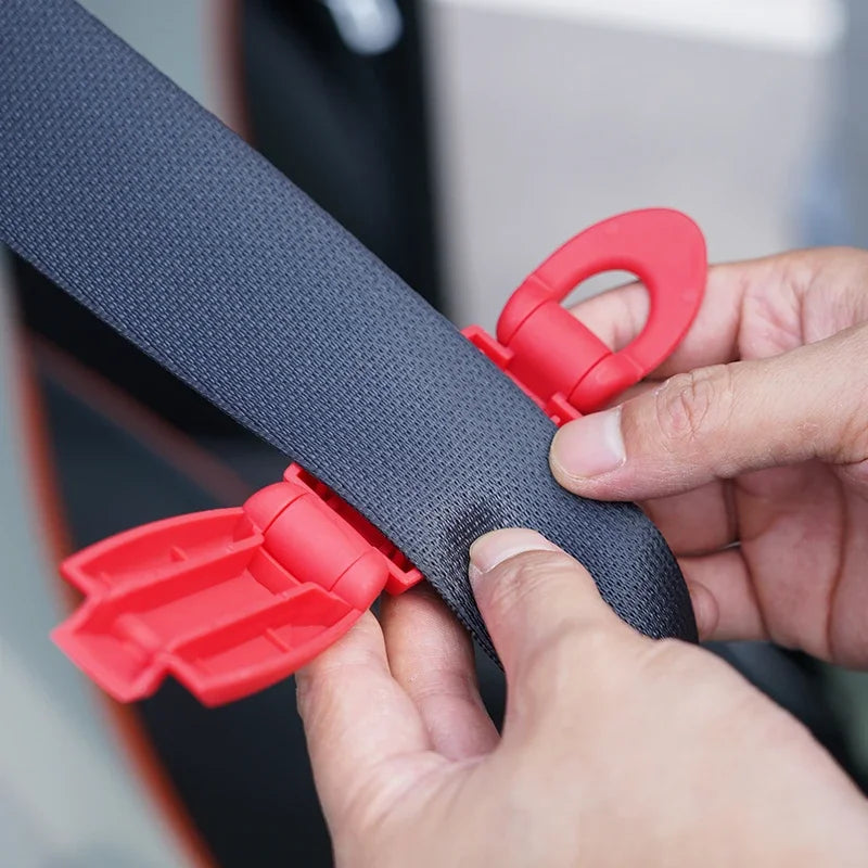 A hand is holding a red seat belt clip, which is attached to a dark gray strap, demonstrating the product.