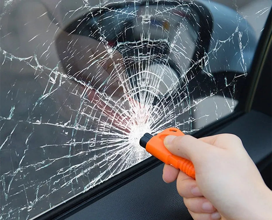 A hand uses an orange tool to break a spiderweb crack in a car window.