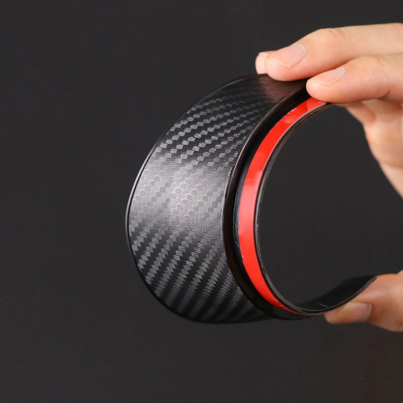 A hand holds a black carbon fiber automotive rearview mirror rain and dust visor.
