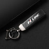 A black carbon fiber style keychain with silver hardware and white lettering is displayed on a dark surface.