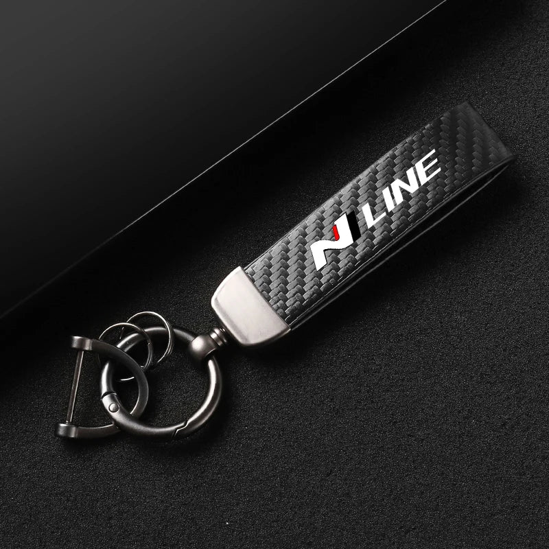 A black carbon fiber style keychain with silver hardware and white lettering is displayed on a dark surface.