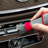 A hand uses a red and gray brush to clean the air vent of a car interior.