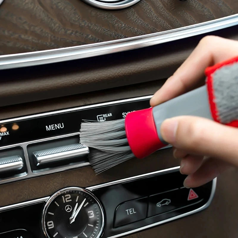 A hand uses a red and gray brush to clean the air vent of a car interior.
