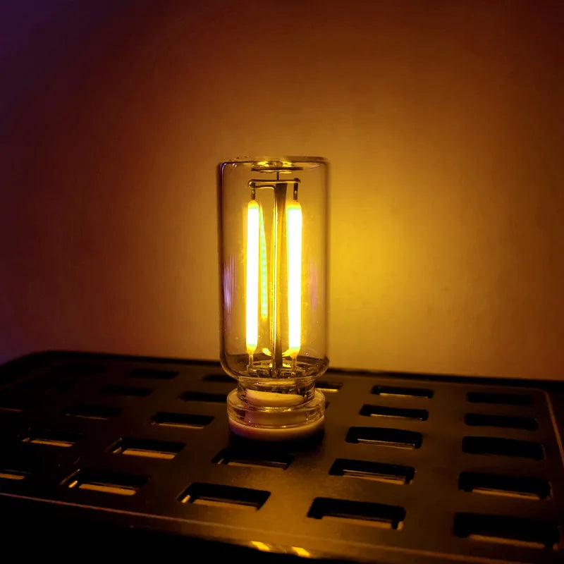 A clear glass LED night light with a warm yellow glow sits on a black metal grate.