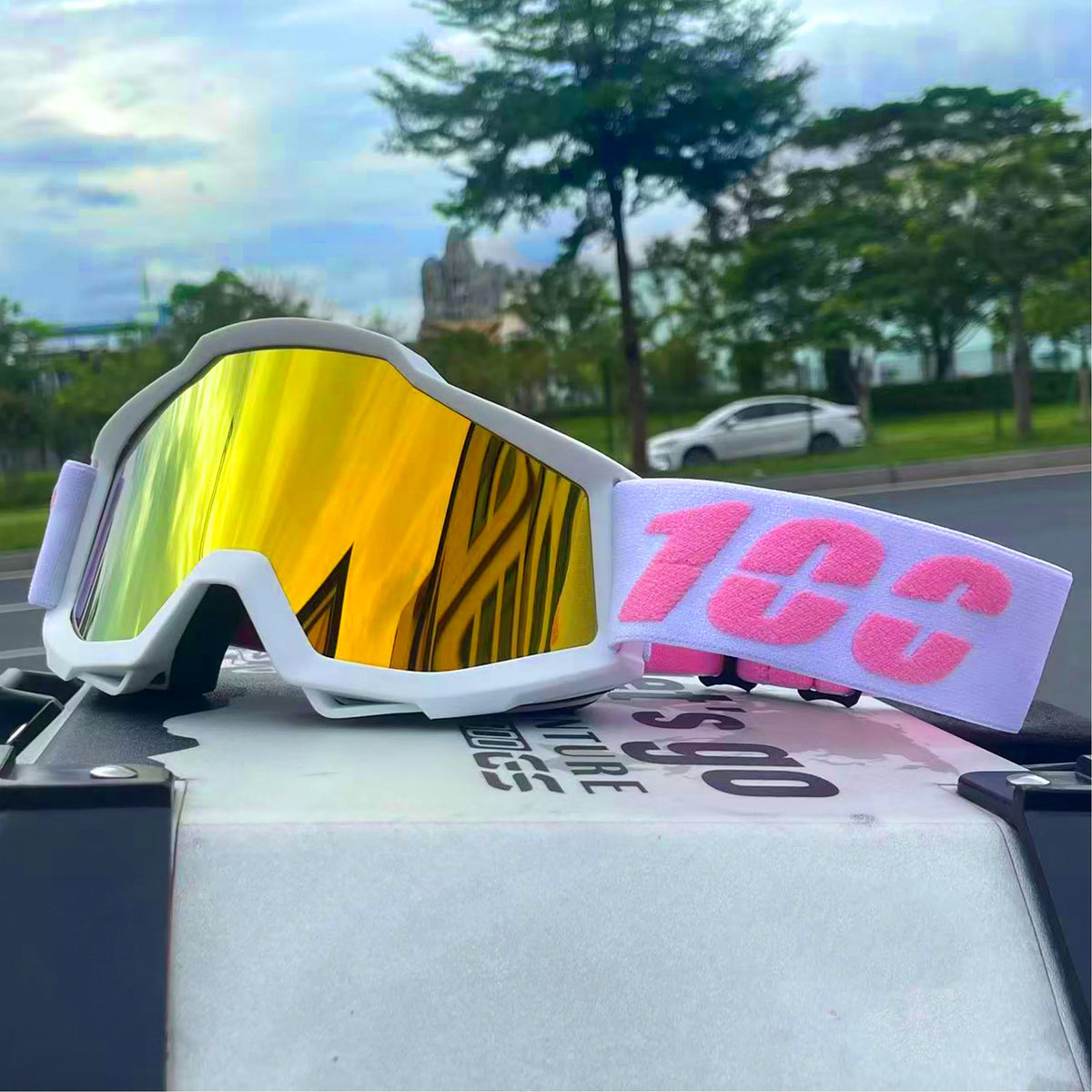 A pair of orange-tinted off-road motorcycle goggles with white frames and pink strap resting on a surface.
