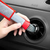 A hand is using a red and gray brush to clean the air vent of a car, near an airbag warning label.