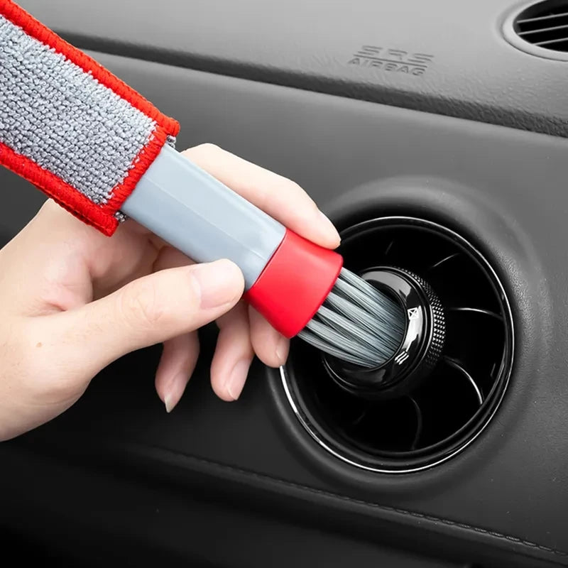 A hand is using a red and gray brush to clean the air vent of a car, near an airbag warning label.
