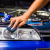 A hand is applying a dark blue microfiber pad to the headlight of a blue car for polishing or waxing.