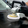 A hand wearing a glove is using a polishing device with a foam disc on a dark car surface.