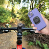 A hand holds a purple universal motorcycle phone mount attached to a bicycle handlebar in a wooded outdoor setting.