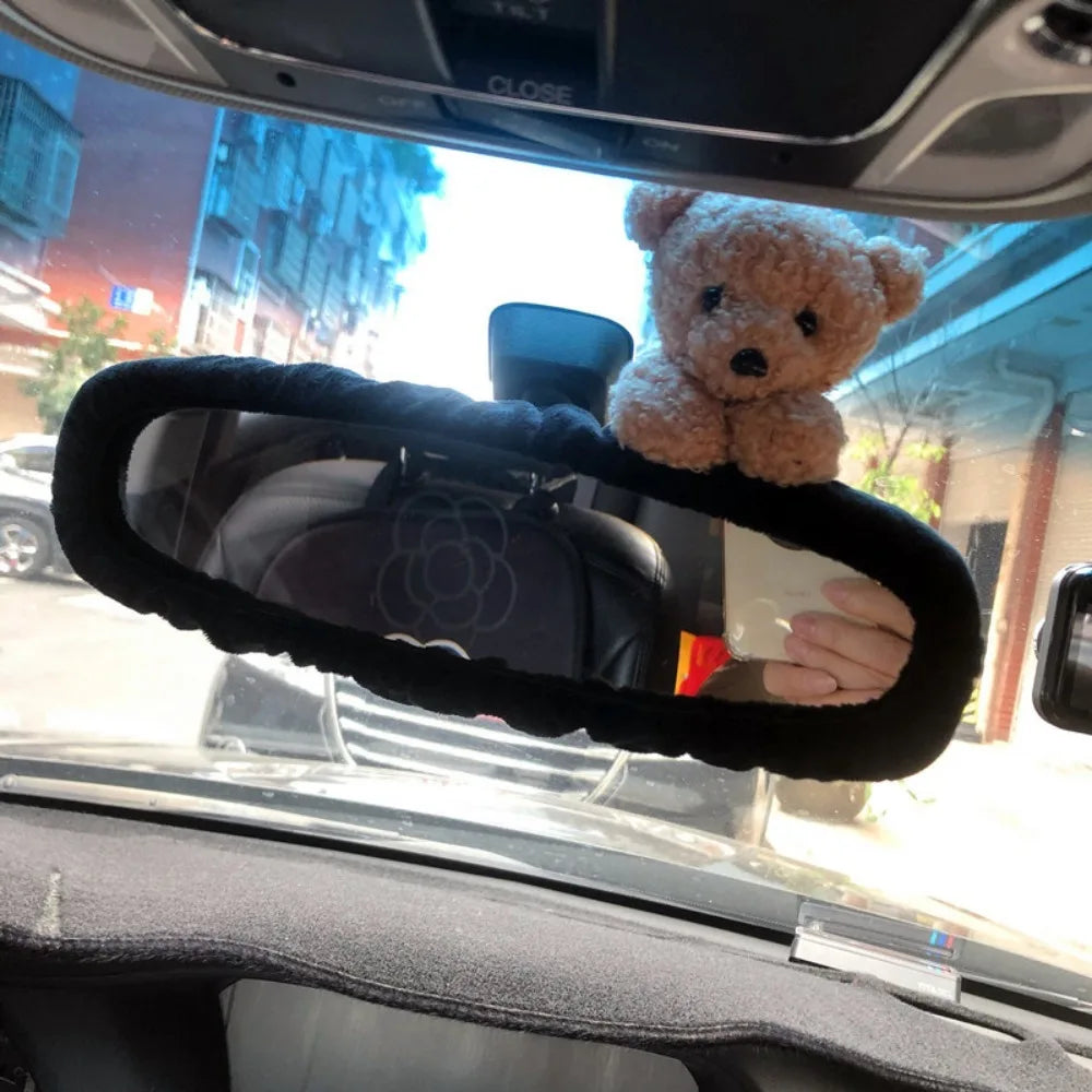 A fluffy brown teddy bear is placed on a black rearview mirror cover inside a car.