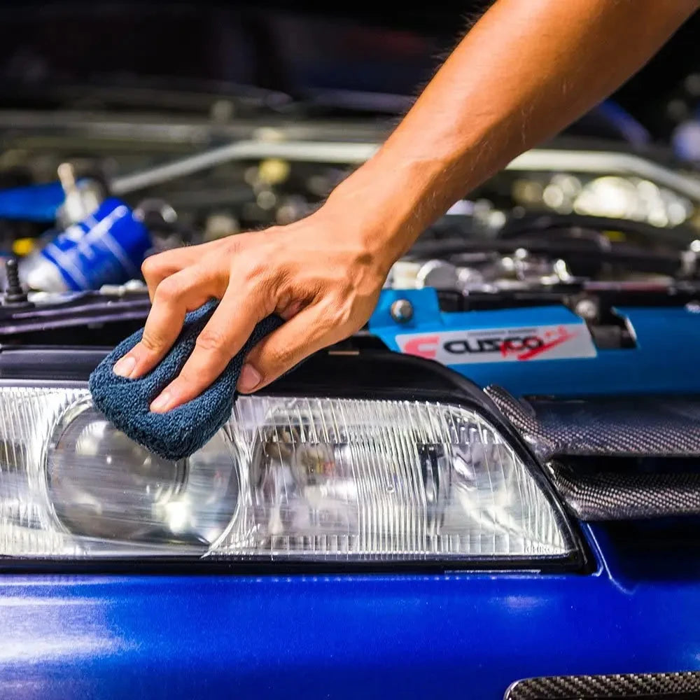 A hand is applying a dark blue microfiber pad to the headlight of a blue car for polishing or waxing.