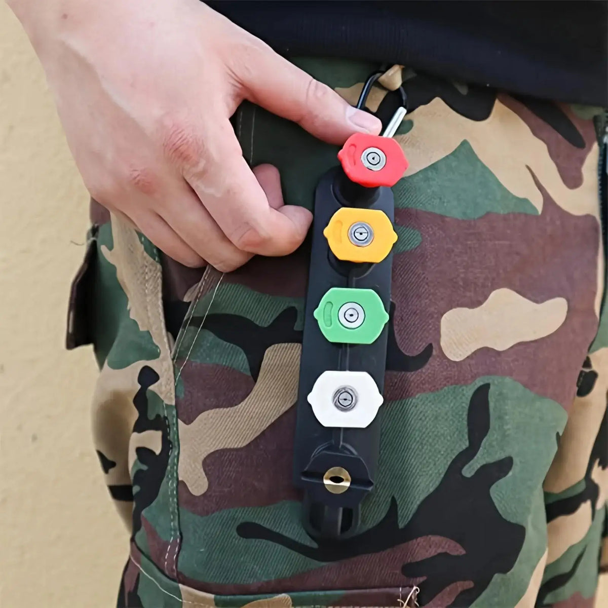 A hand holds a black high-pressure water gun with colored nozzles against a camouflage fabric.
