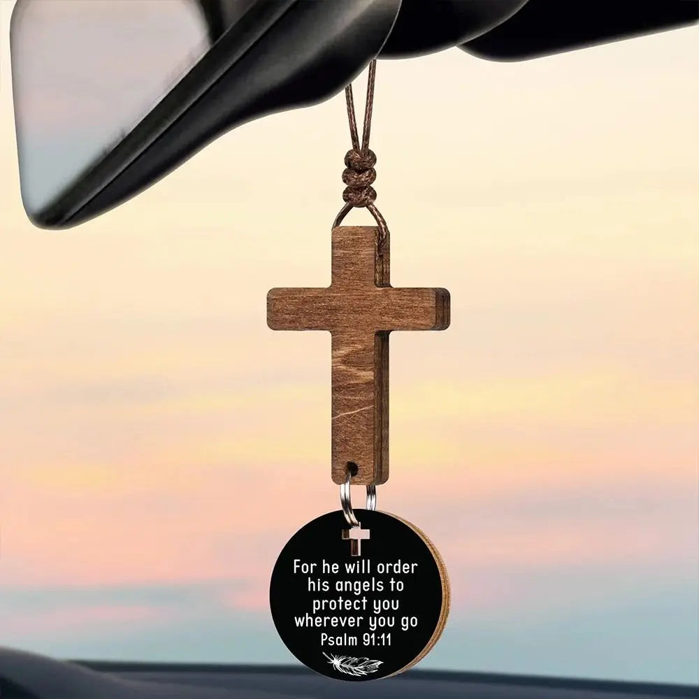 A wooden cross ornament hangs from a black object, featuring the text For he will order his angels to protect you wherever yo