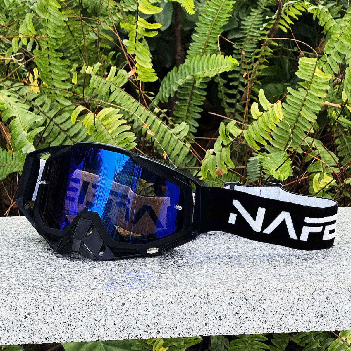 Eine schwarze und blaue Motocross-Schutzbrille liegt auf einer hellgrauen Oberfläche mit grünem Laub im Hintergrund.