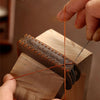 A hand is using a thin orange leather cord to attach a leather key fob cover to a wooden block.