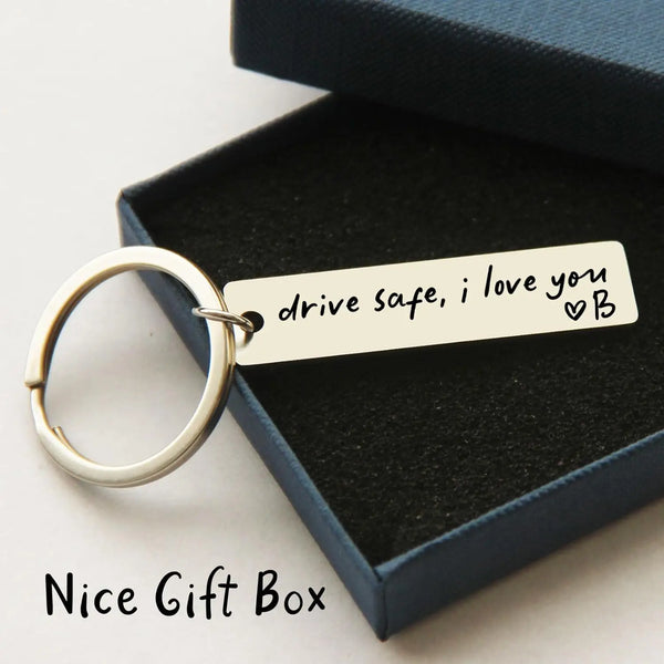 A silver keychain with a custom engraved tag reading drive safe i love you and a heart symbol inside a dark blue gift box.