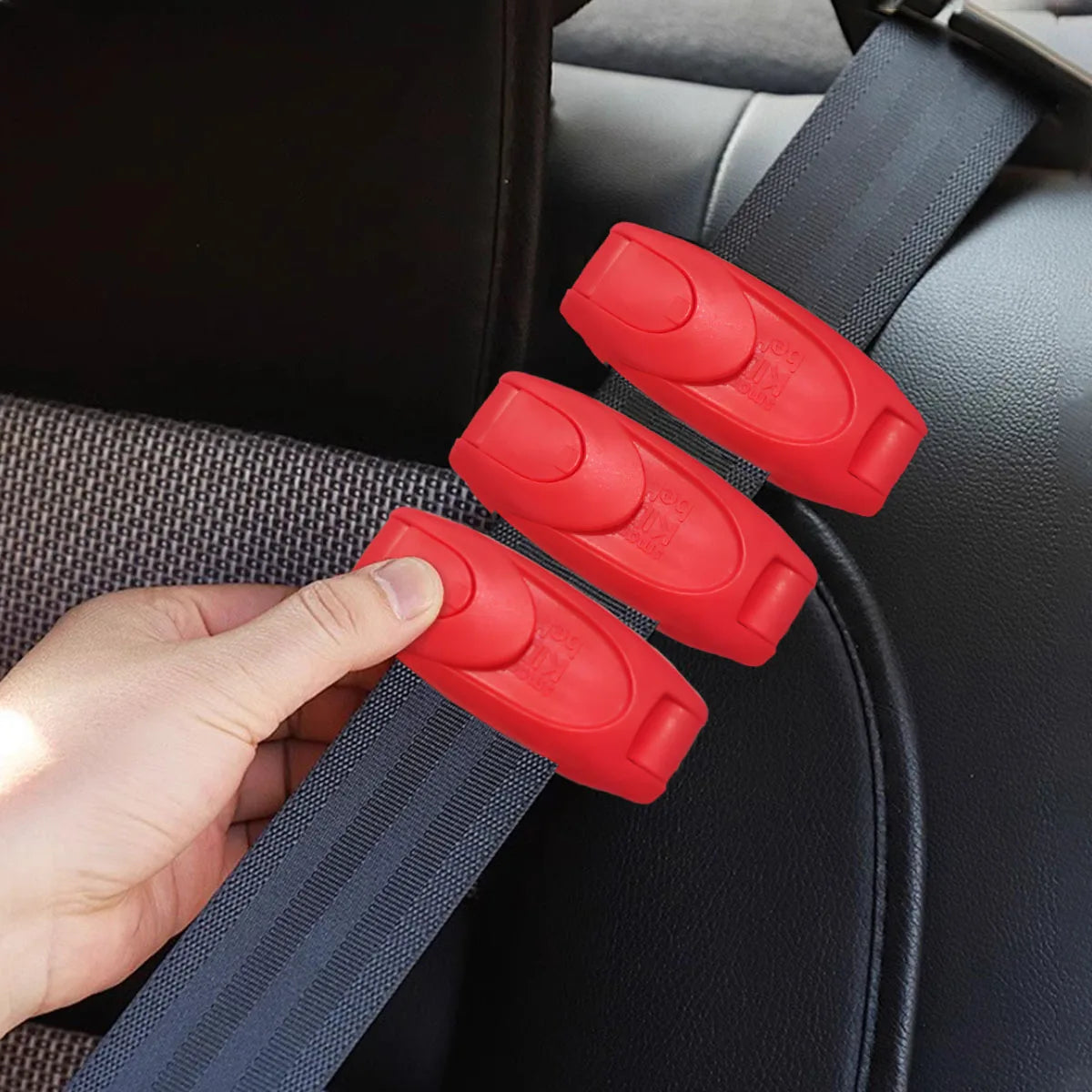 A hand is holding red comfort-adjustable seat belt clips attached to a dark seat belt strap.